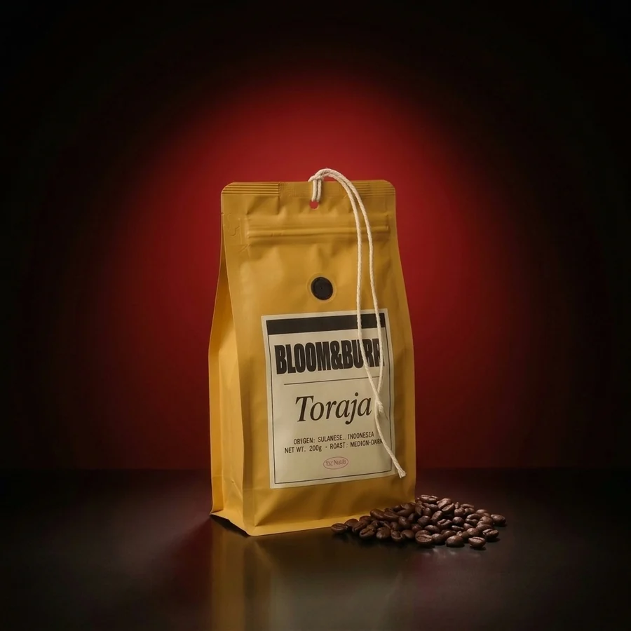 The Toraja pouch standing on the glossy floor in the same glow, a heap of roasted beans beside it